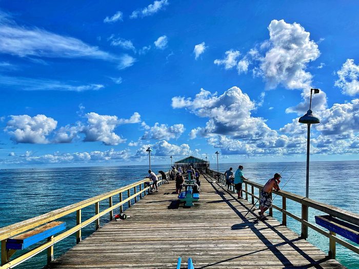 A long wooden fishing pier running out over bright blue water under a clouded sky.