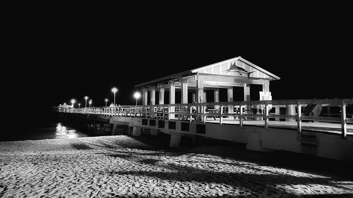 Black-and-white night photo of the pier and its lit pavilion over dark sand.
