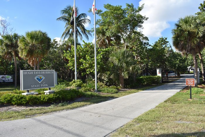 A park entrance sign under tall palms beside a paved path.