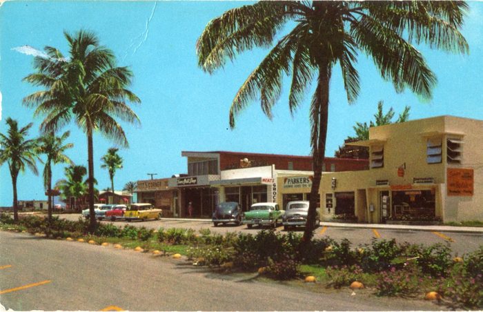 Vintage color photo of a low shopping center with palm trees and 1950s cars near the beach.
