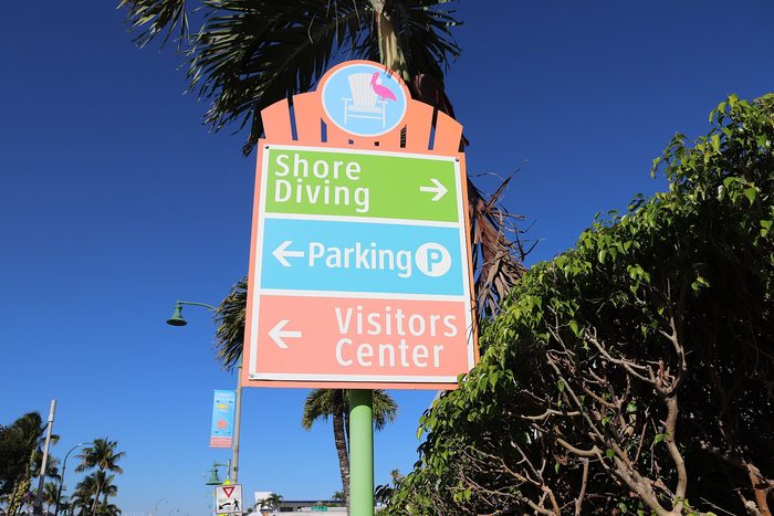 A colorful wayfinding sign pointing to shore diving, parking and the visitors center, under palm fronds.